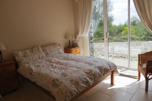 Une chambre à coucher comprenant un lit, une table de chevet, une lampe, un fauteuil en osier et une vue sur un jardin, visible à travers une porte vitrée.