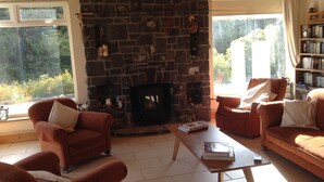 A cozy living room with a stone fireplace, comfortable seating, and large windows offering a view of greenery. - Lakeside Country Cottage. 
 (Headford)