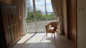 A room with a sliding glass door leading to a stone pathway and greenery. There is a wicker chair and a wooden wardrobe. - Lakeside Country Cottage. 
 (Headford)