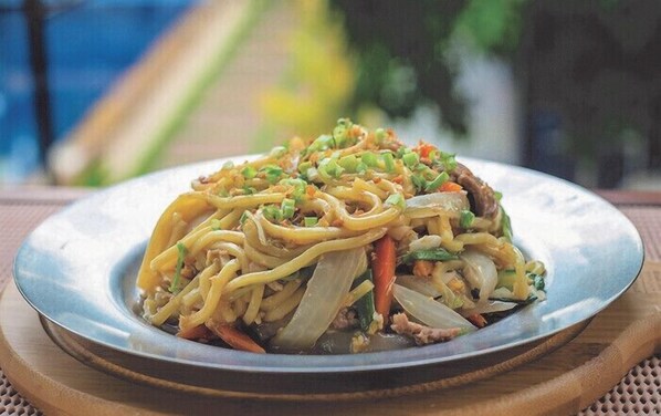 A plate of noodles with vegetables and meat, served on a metal plate with a wooden placemat underneath.