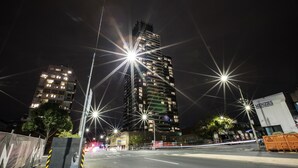 Paisagem urbana noturna com um edifício alto, com várias varandas e janelas iluminadas. Faróis de rua e veículos criam efeitos de luzes cintilantes.