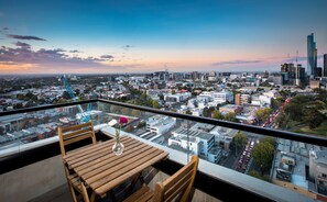 A cityscape view from a high-rise balcony with a wooden table and chairs, a vase with flowers, and a clear glass railing. - Spencer Street Apartments (West Melbourne)