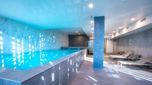 Piscina interna com água cristalina, cercada por espreguiçadeiras e um design moderno com paredes e luminárias de mármore.