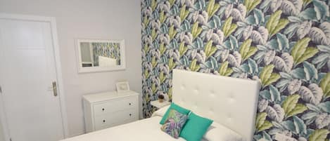 A bedroom with a white bed, a nightstand, a dresser, a mirror, and a wall with a leafy pattern.