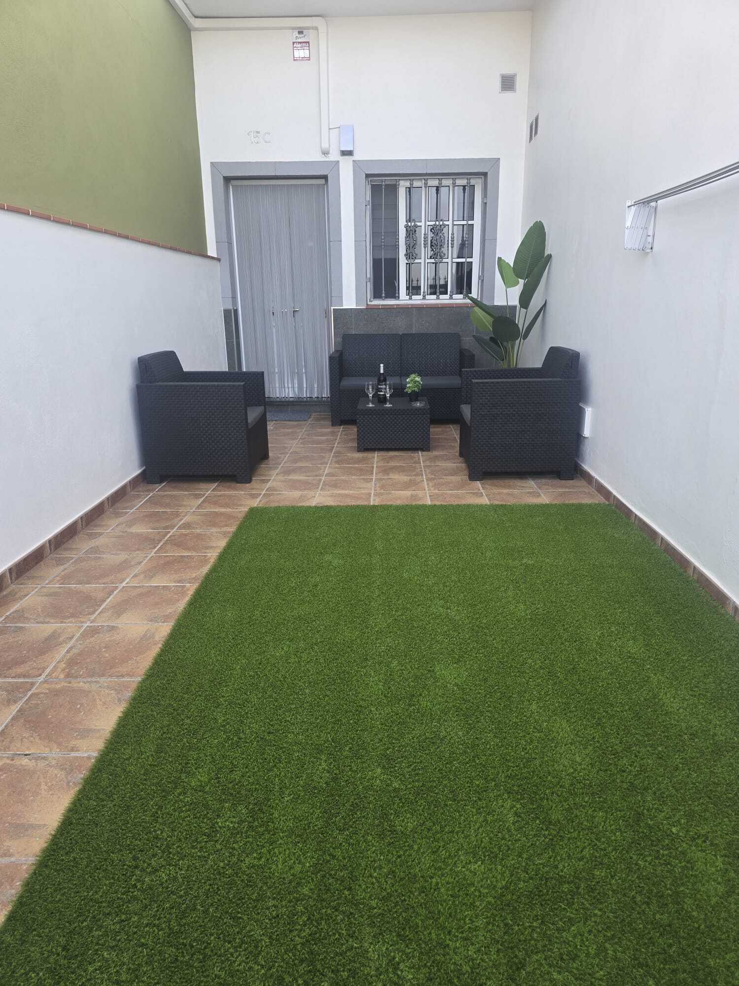 A courtyard with a small artificial grass area, a seating arrangement with sofas and a coffee table, and a door with a window and decorative ironwork.