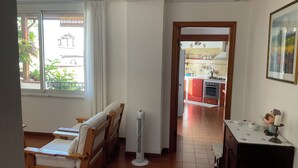 Una habitación con una cama de madera, un ventilador de pie y vista a una cocina por una puerta abierta.
