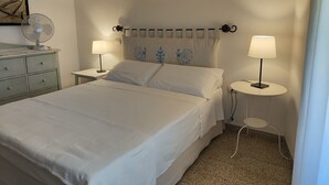 Una cama bien hecha con sábanas blancas, dos mesitas de noche con lámparas, un ventilador de pared y un cabecero decorativo.