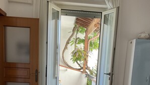 Una habitación con puerta de madera, cama con colcha azul, una silla de mimbre y vista a un balcón con un árbol y piso de baldosas.