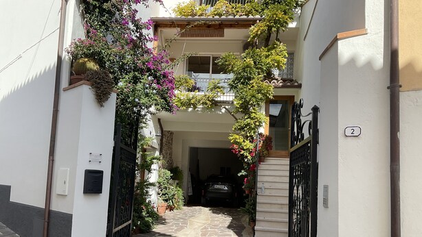 Un estrecho callejón empedrado que conduce a un edificio con balcón, rodeado de macetas con plantas y flores.