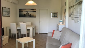 A living room with a grey sofa, a small white table, and a dining area with chairs and a table. There is a painting on the wall and an air conditioning unit above the door.