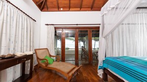 Una habitación con piso de madera, una cama con colcha azul, una silla de mimbre con almohada verde, una mesa de madera con un libro y una bandeja, y vista a la playa a través de una puerta de vidrio.