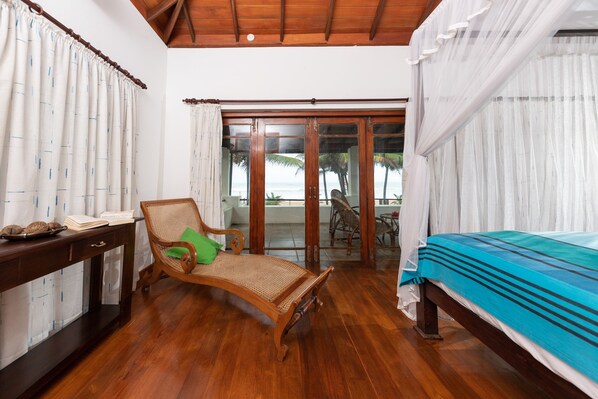 A room with a wooden floor, a bed with a blue bedspread, a wicker chair with a green pillow, a wooden table with a book and a tray, and a view of the beach through a glass door.