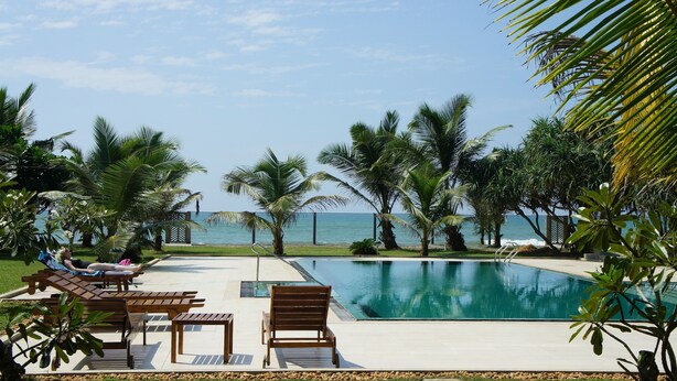 Zona de piscina con tumbonas y sillones de madera, rodeada de palmeras y vista al mar.