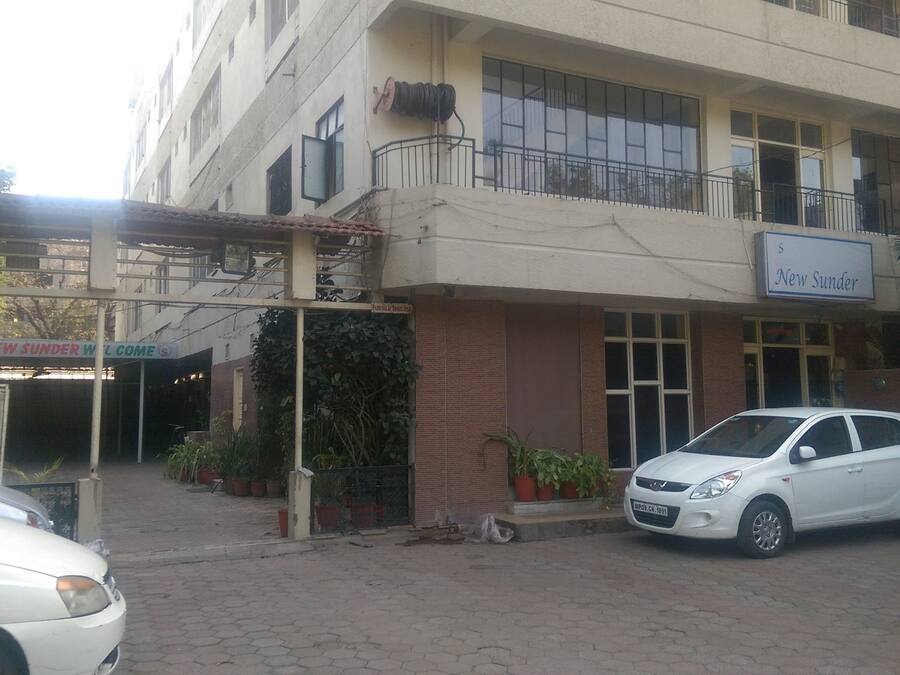 Ein Hotel Eingang mit einem Schild, auf dem 'Hotel New Sunder Willkommen' steht. Vor dem Gebäude sind Autos geparkt, und es hat mehrere Fenster und Balkone.