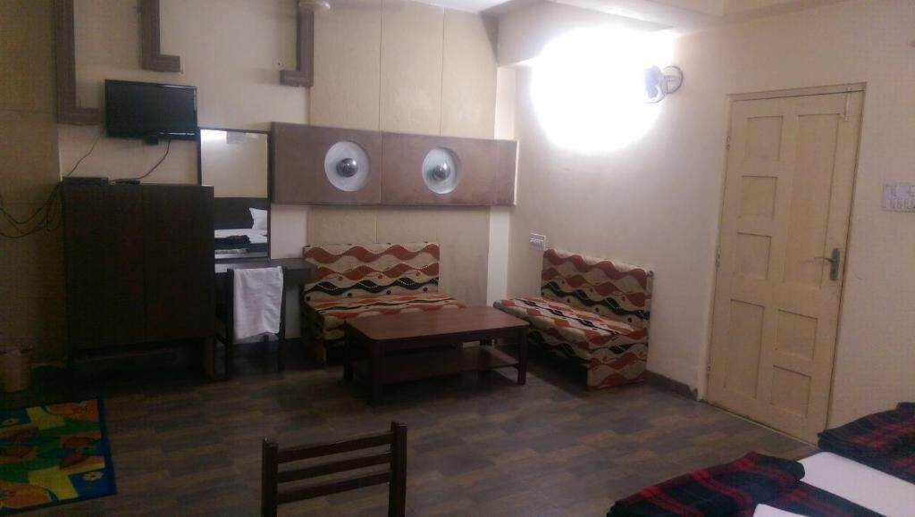 Una habitación con cama, escritorio, silla y mesa. Hay un televisor en la pared y una puerta con manija.
