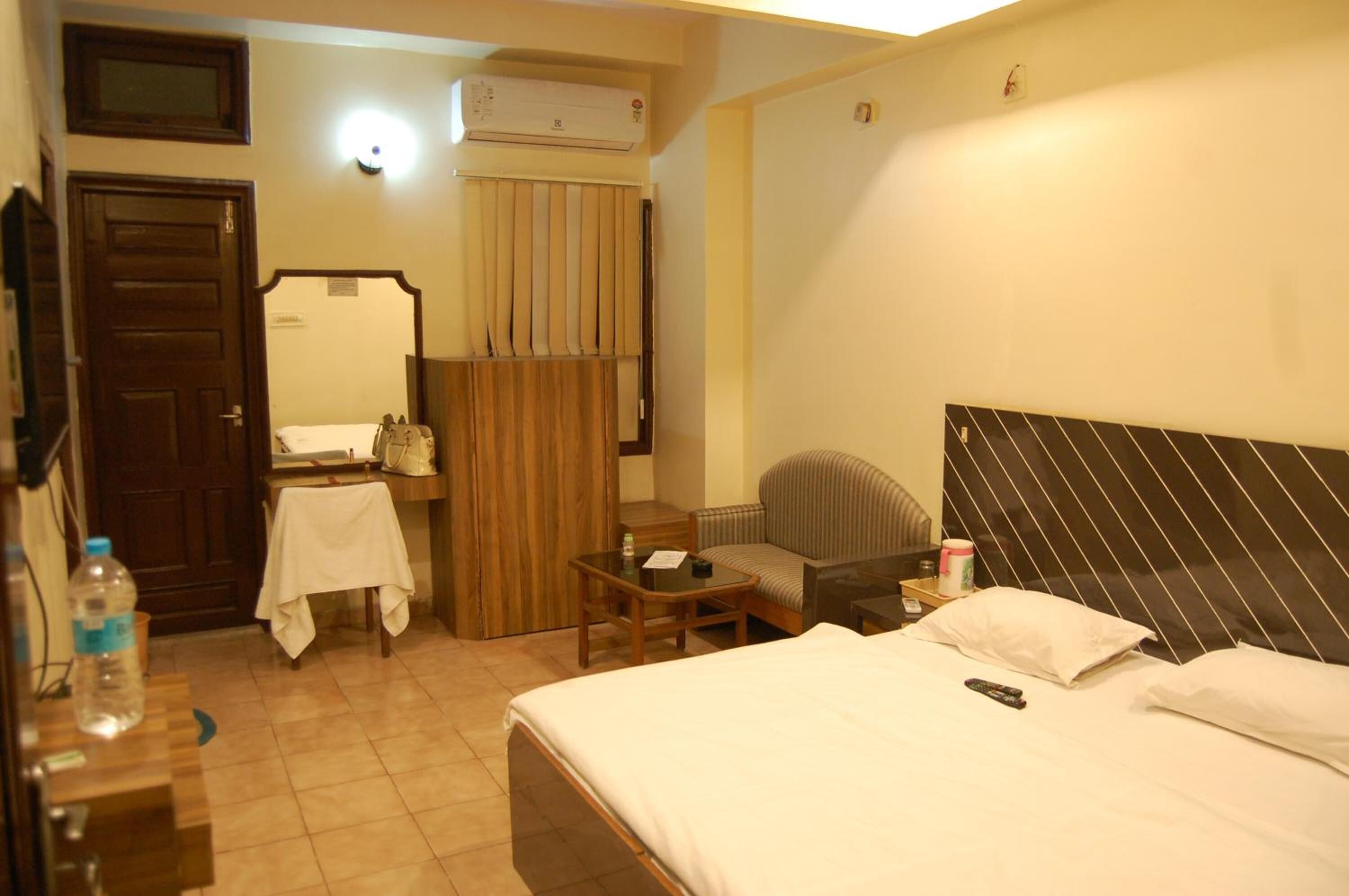 A hotel room with a bed, a chair, a small table, a mirror, a wardrobe, and an air conditioning unit.