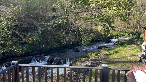 A wooden deck overlooking a flowing stream with a rustic cabin in the background. - Riverside 2 Bed Lodge with Hot Tub. Walk to Lynton Centre. Free Parking. (Lynton)