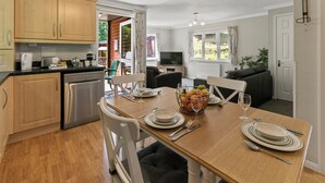 A modern kitchen with wooden cabinets, a dining table set for four, and a cozy living area with a sofa and TV. - Riverside 2 Bed Lodge with Hot Tub. Walk to Lynton Centre. Free Parking. (Lynton)