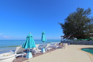 A poolside area with lounge chairs and umbrellas, a clear view of the ocean, and a tree in the background.
