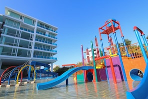 A water park with colorful slides and play structures, a modern building with balconies, and a clear blue sky.