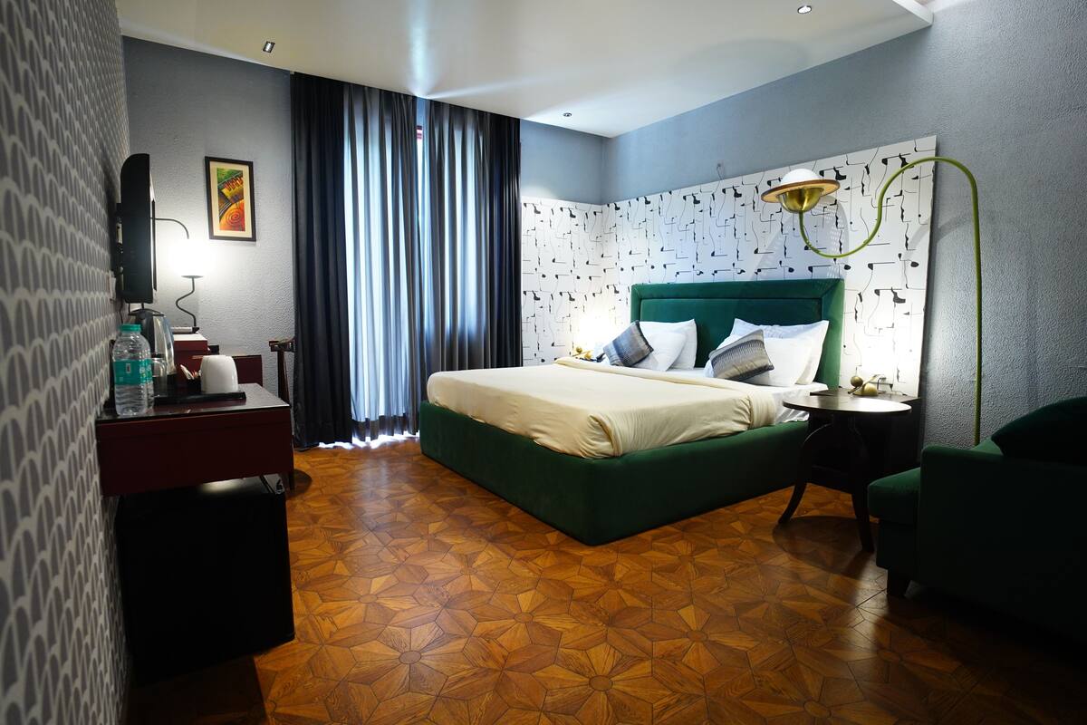 A modern hotel room with a green upholstered bed, a flat-screen TV, a desk with a lamp, and a patterned wall.