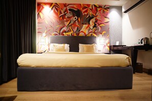 A large bed with a dark headboard, a colorful abstract mural on the wall behind, and a desk with a kettle and a cup on it. -  The Grand Vikalp by Saga Hotels (New Delhi)
