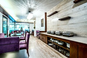 A modern dining area with purple chairs, a buffet counter with food containers, and a view of the dining room through glass doors. -  The Grand Vikalp by Saga Hotels (New Delhi)