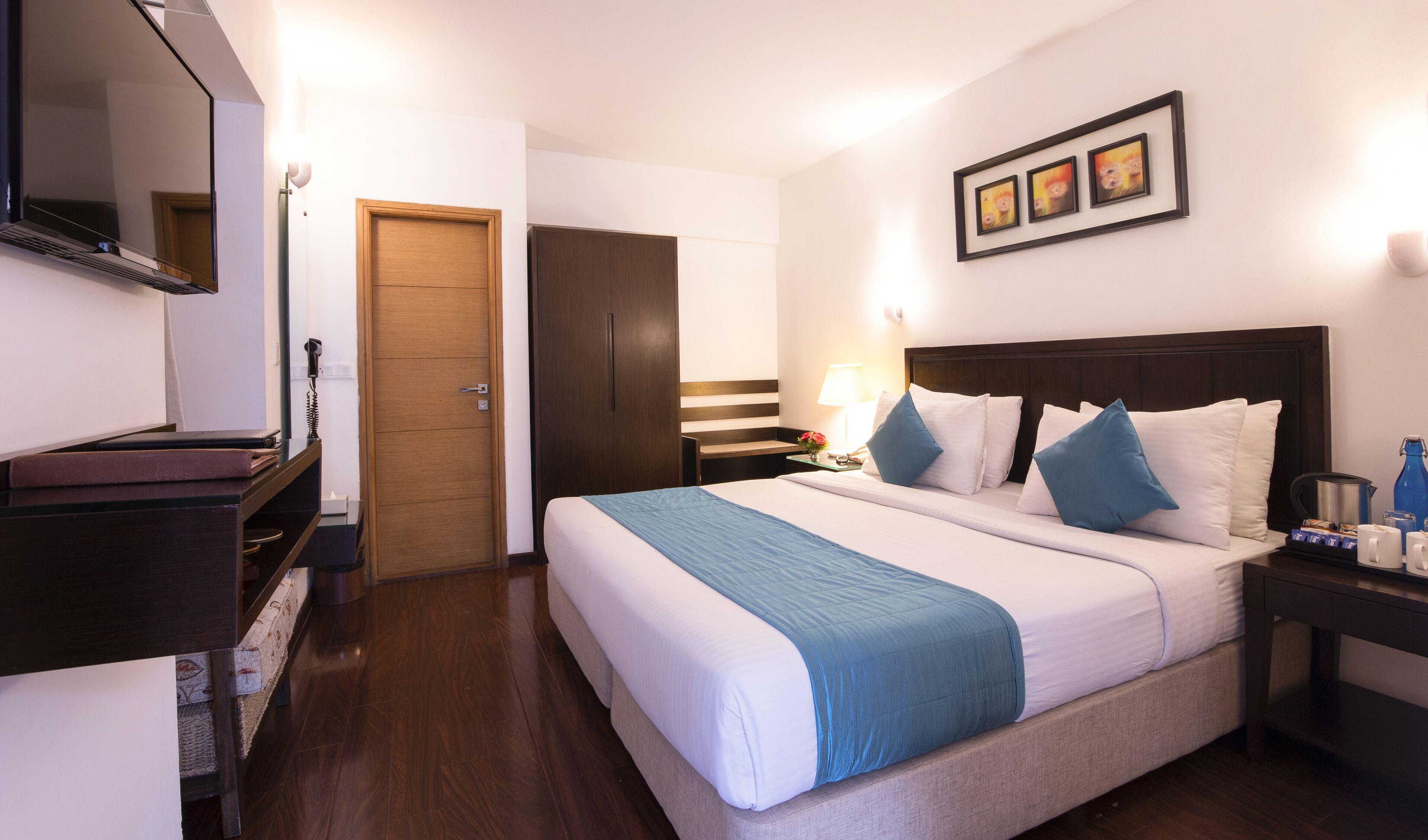 A hotel room with a large bed, a wooden wardrobe, a desk with a lamp, and a framed picture on the wall.