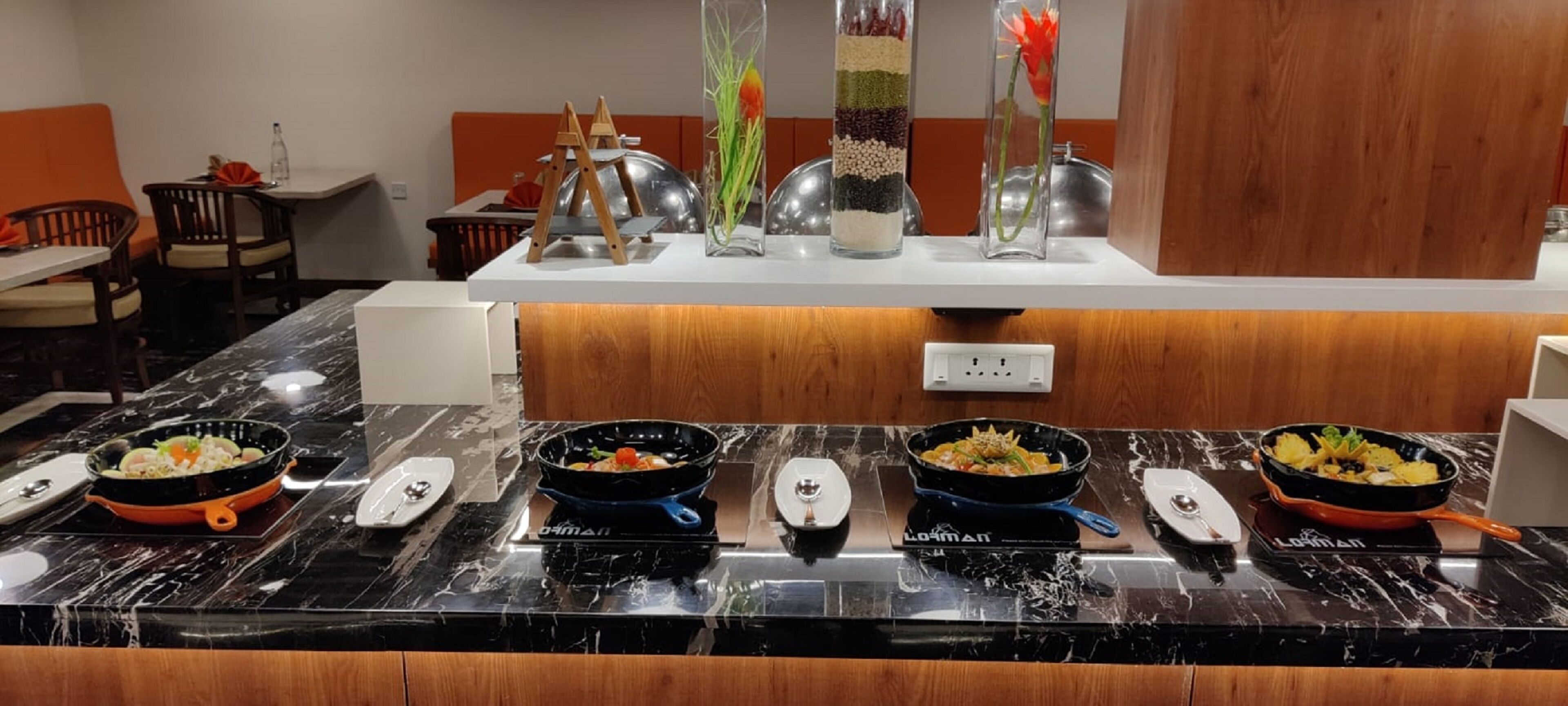 Un restaurante con mostrador de mármol que exhibe diferentes platos, pared de paneles de madera y una zona de estar con cojines anaranjados.