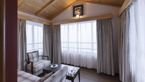 Una habitación con techo de madera, ventanas amplias con cortinas, un cuadro enmarcado y una mesa con una silla.