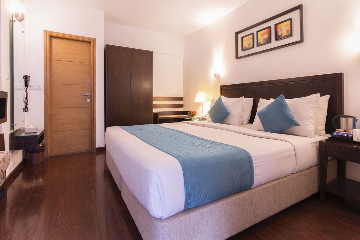 Una habitación de hotel con una cama grande, una mesita de noche con lámpara, una mesita auxiliar y un cuadro colgado en la pared.