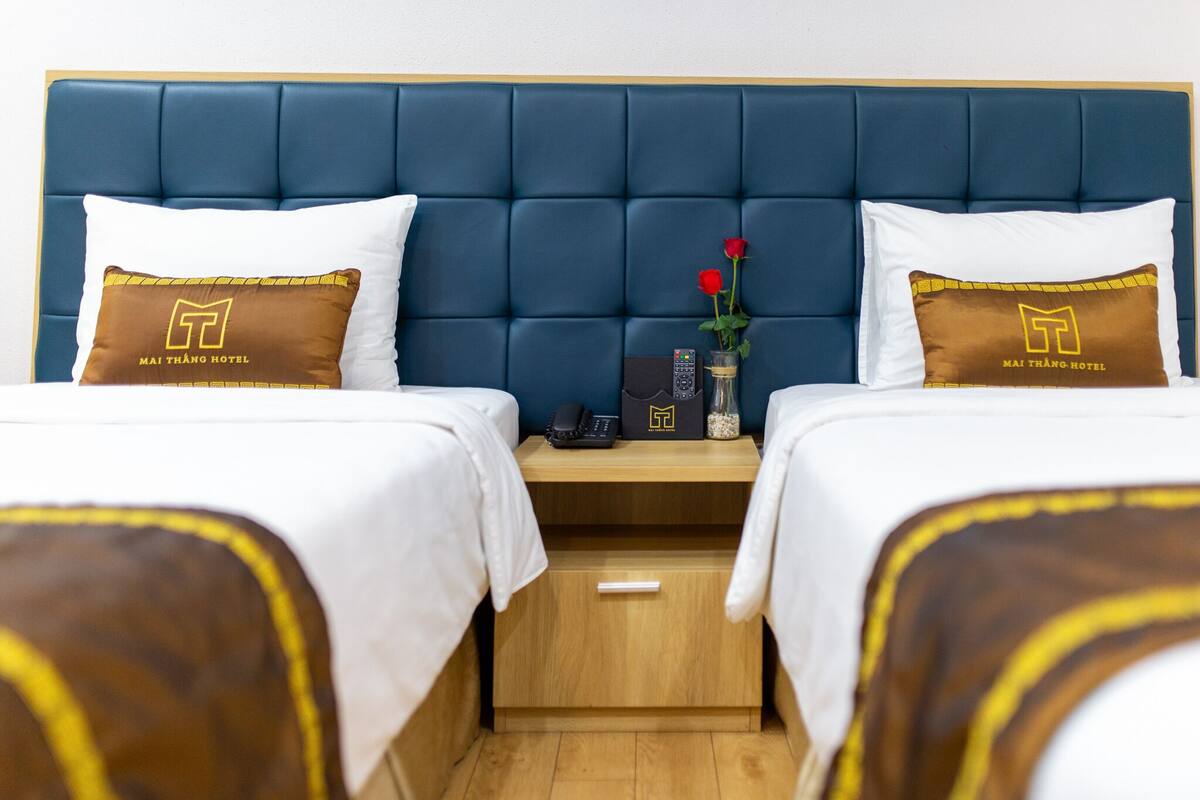 Habitación de hotel con dos camas, cada una con un almohadón que muestra el logotipo del hotel. Se observa una mesita de noche con un teléfono y un jarrón con flores.