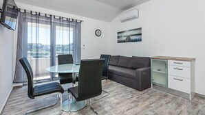 A modern living room with a glass dining table, black chairs, a grey sofa, a small kitchen area with a white cabinet, and a large window with sheer curtains. - 2 bedroom amazing apartment in Split (Split)