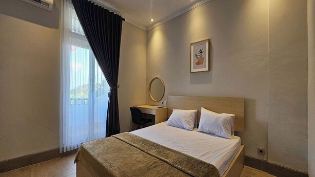 A hotel room with a bed, a desk, a chair, a round mirror, and a window with curtains. - Hotel Bugis Asri (Yogyakarta)