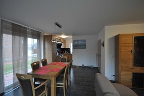 A dining area with a wooden table and chairs, a kitchen in the background, and a view of the outdoors through large windows.