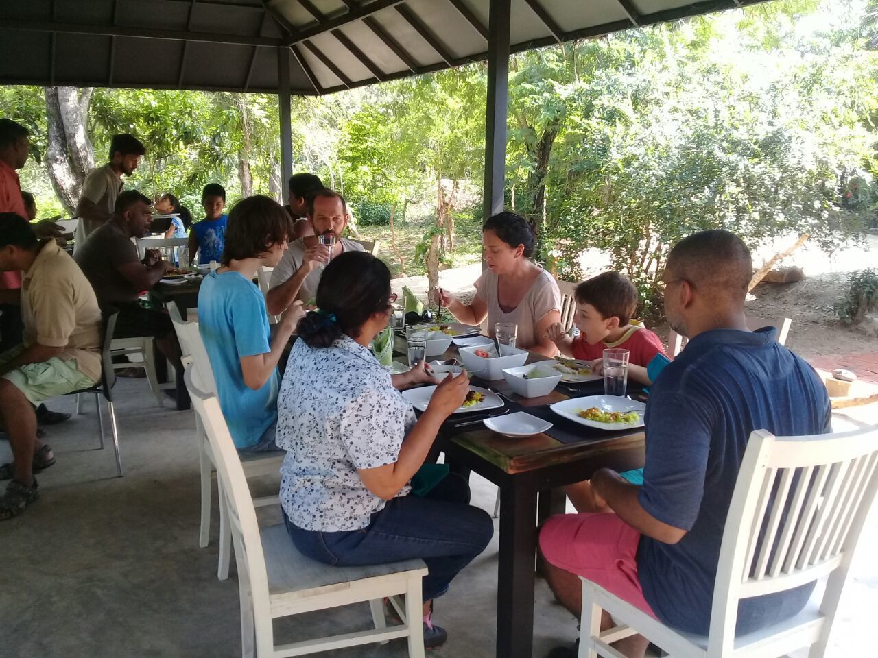 A covered outdoor dining area with people seated at tables, surrounded by greenery.