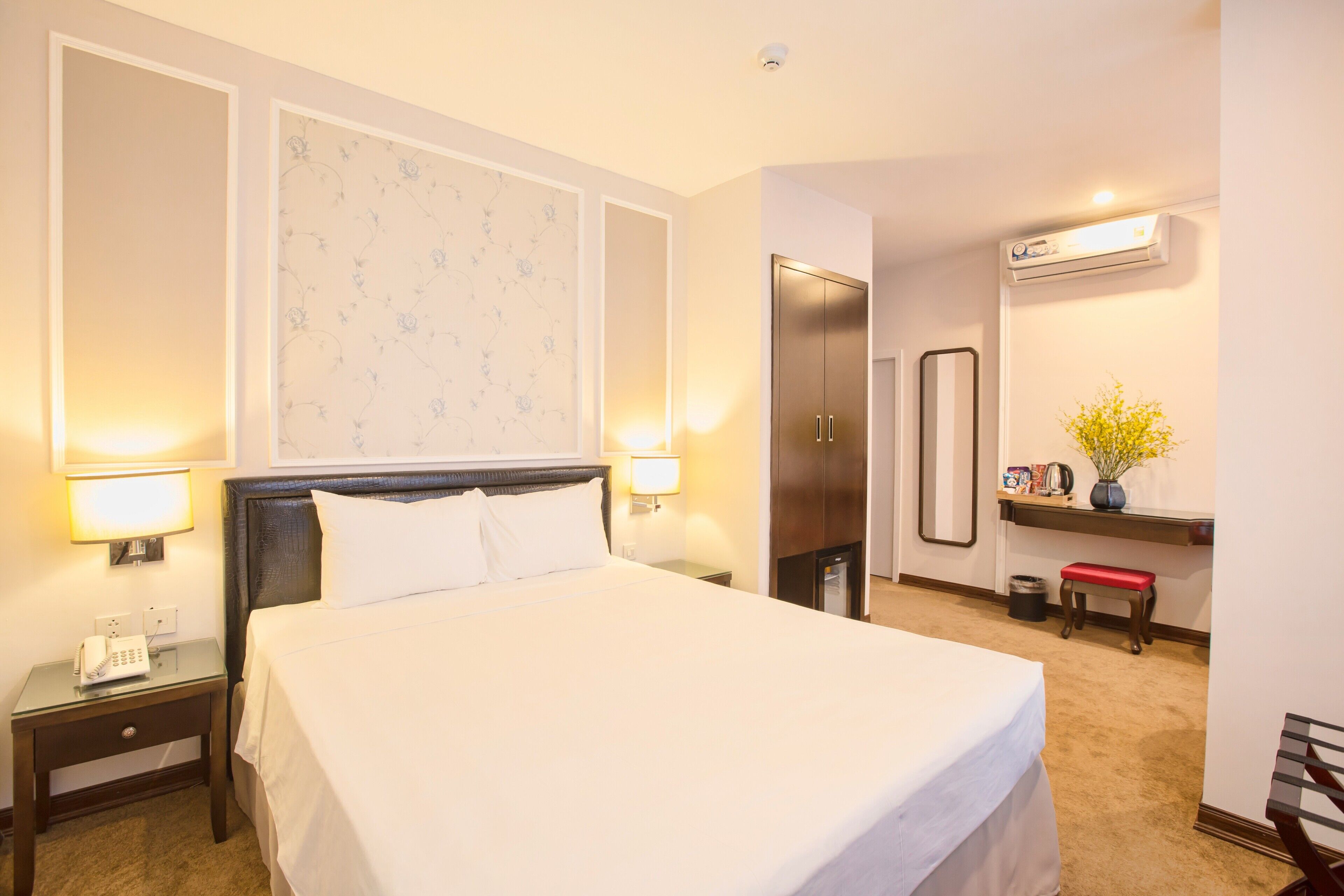 Ein Hotelzimmer mit einem großen Bett, Nachttischlampen, einem kleinen Tisch mit Telefon, einem Spiegel, einem Ablagefach für Toilettenartikel und einer Klimaanlage.
