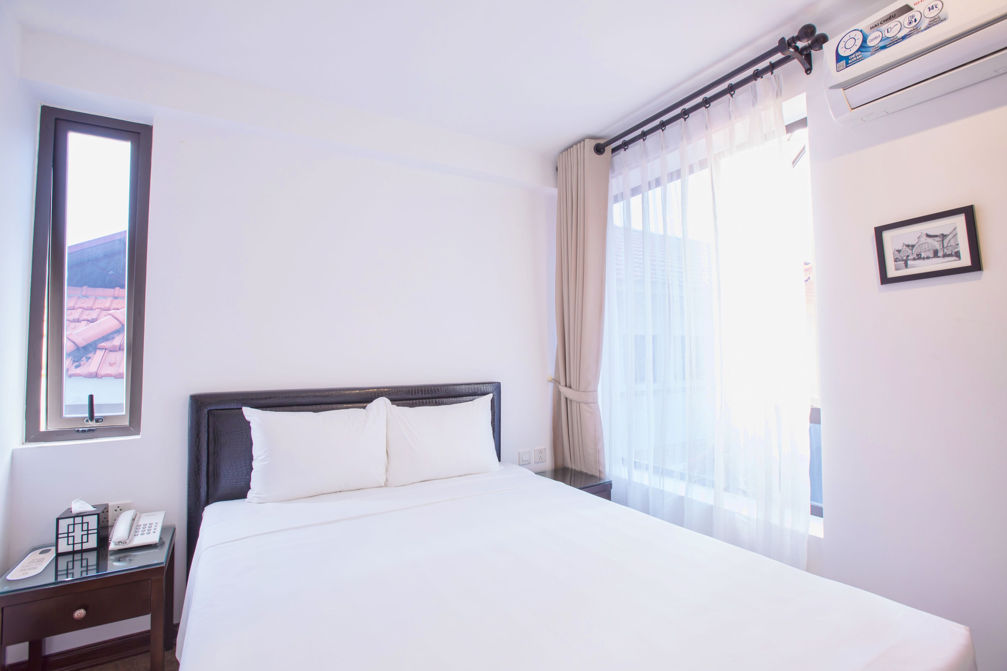 A hotel room with a bed, a bedside table with a telephone, a window with curtains, and a wall-mounted air conditioning unit.