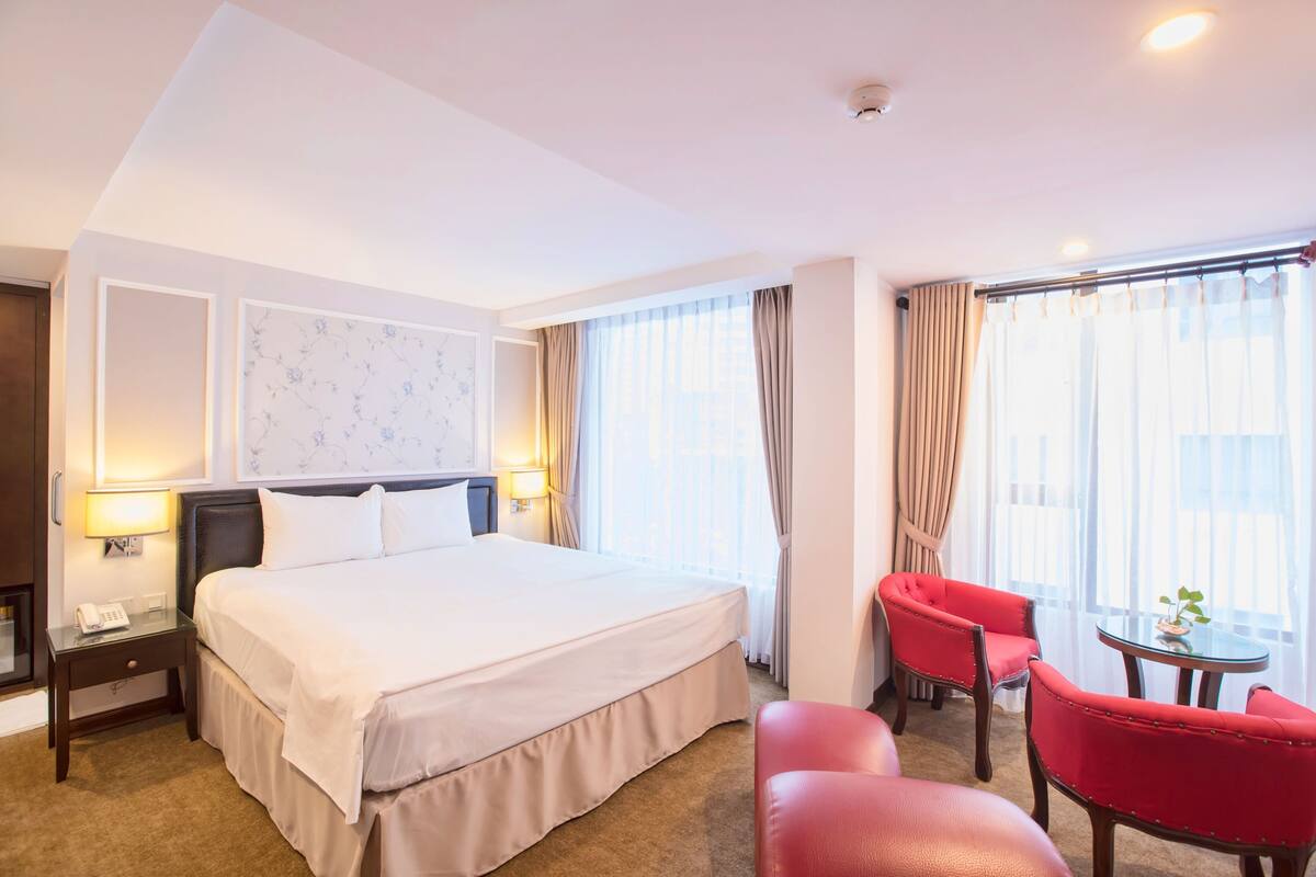 Une chambre d’hôtel avec un grand lit, une table de chevet avec une lampe, une chaise, une petite table avec une plante et une fenêtre avec des rideaux.