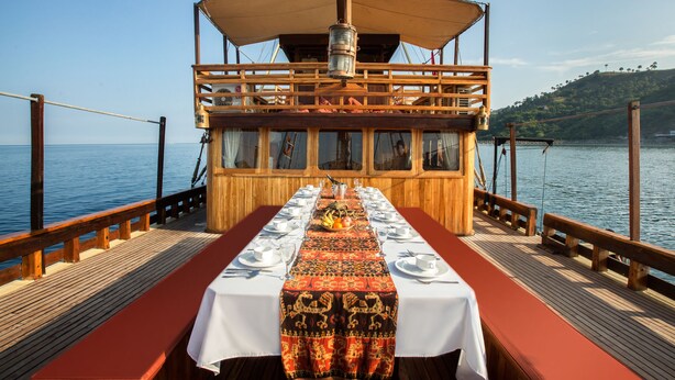 A long table set for dining on a boat with a view of the water and a hilly landscape in the background. - Plataran Phinisi Felicia (Labuan Bajo)