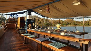 A dining area on a boat with a long wooden table set for a meal, surrounded by a covered deck with a view of the water and hills. - Plataran Phinisi Felicia (Labuan Bajo)