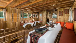 A wooden cabin interior with a dining area, a long table with a black bowl centerpiece, and a sofa with orange cushions. - Plataran Phinisi Felicia (Labuan Bajo)