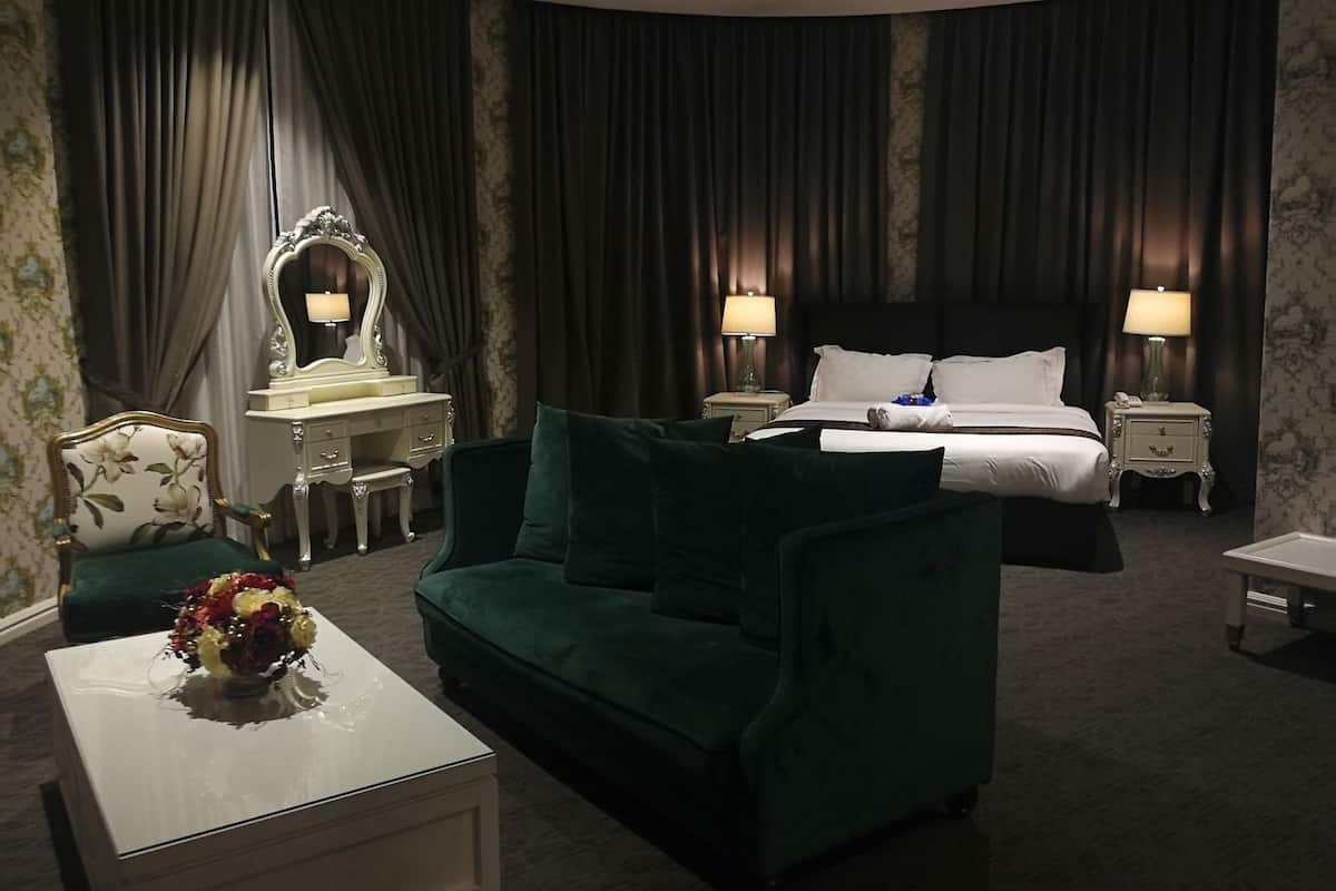 A hotel room with a bed, two bedside tables with lamps, a green sofa, a white desk with a mirror, and a floral vase on a white table.