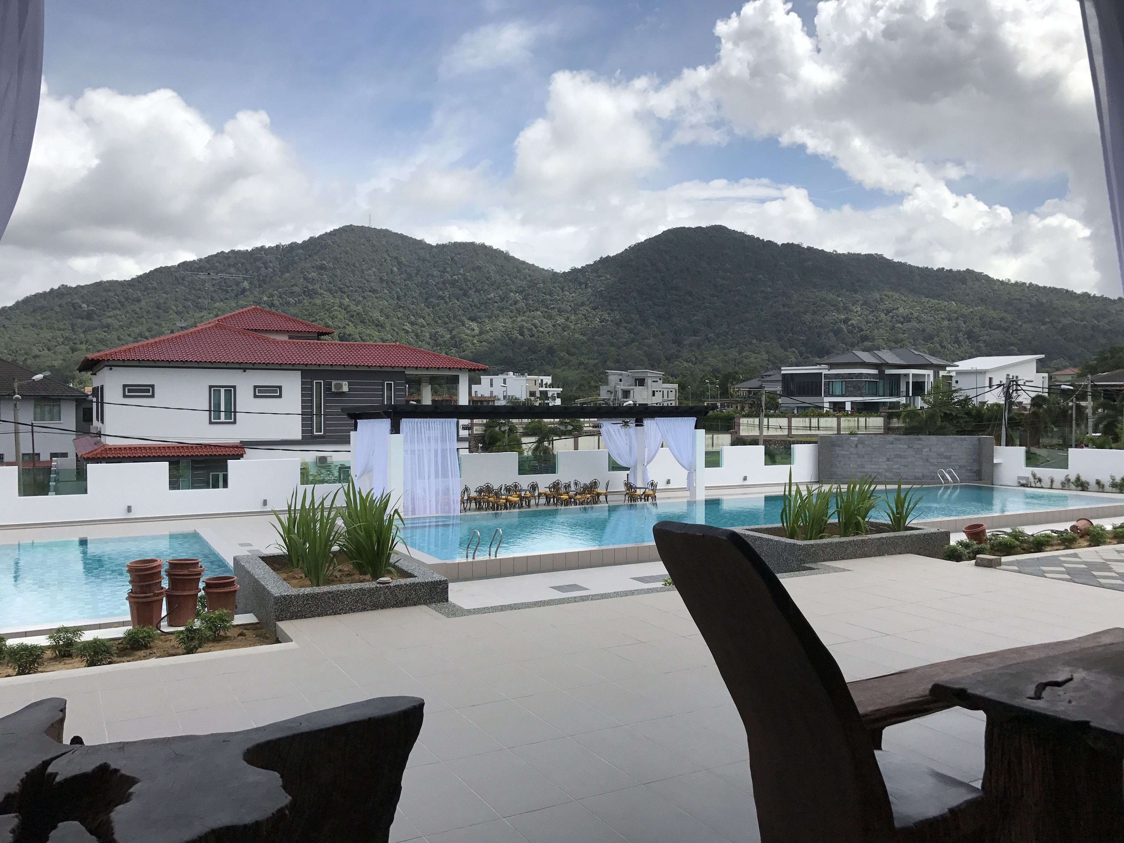 Zona de piscina con vistas a montañas, un edificio blanco de techo rojo y una casa moderna al fondo.