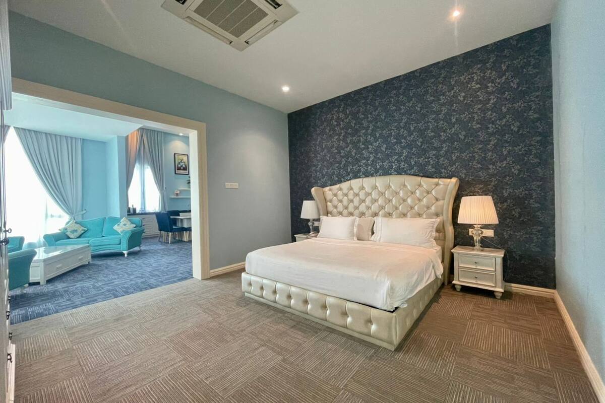 A bedroom with a large bed, a nightstand, a lamp, a dresser, and a view of a living area with a sofa and curtains.