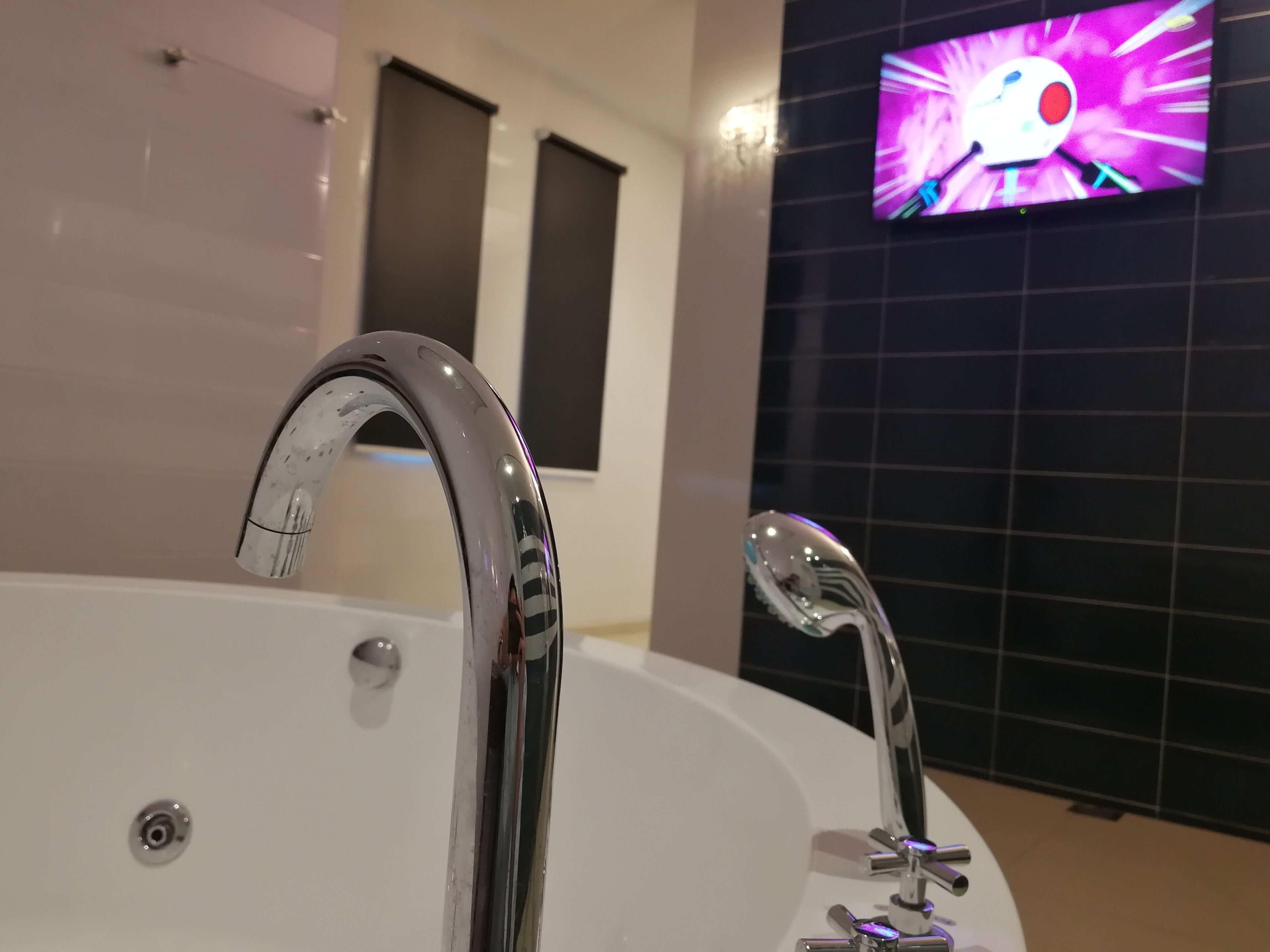A modern bathroom with a large bathtub, chrome fixtures, and a wall-mounted TV displaying a colorful animation.