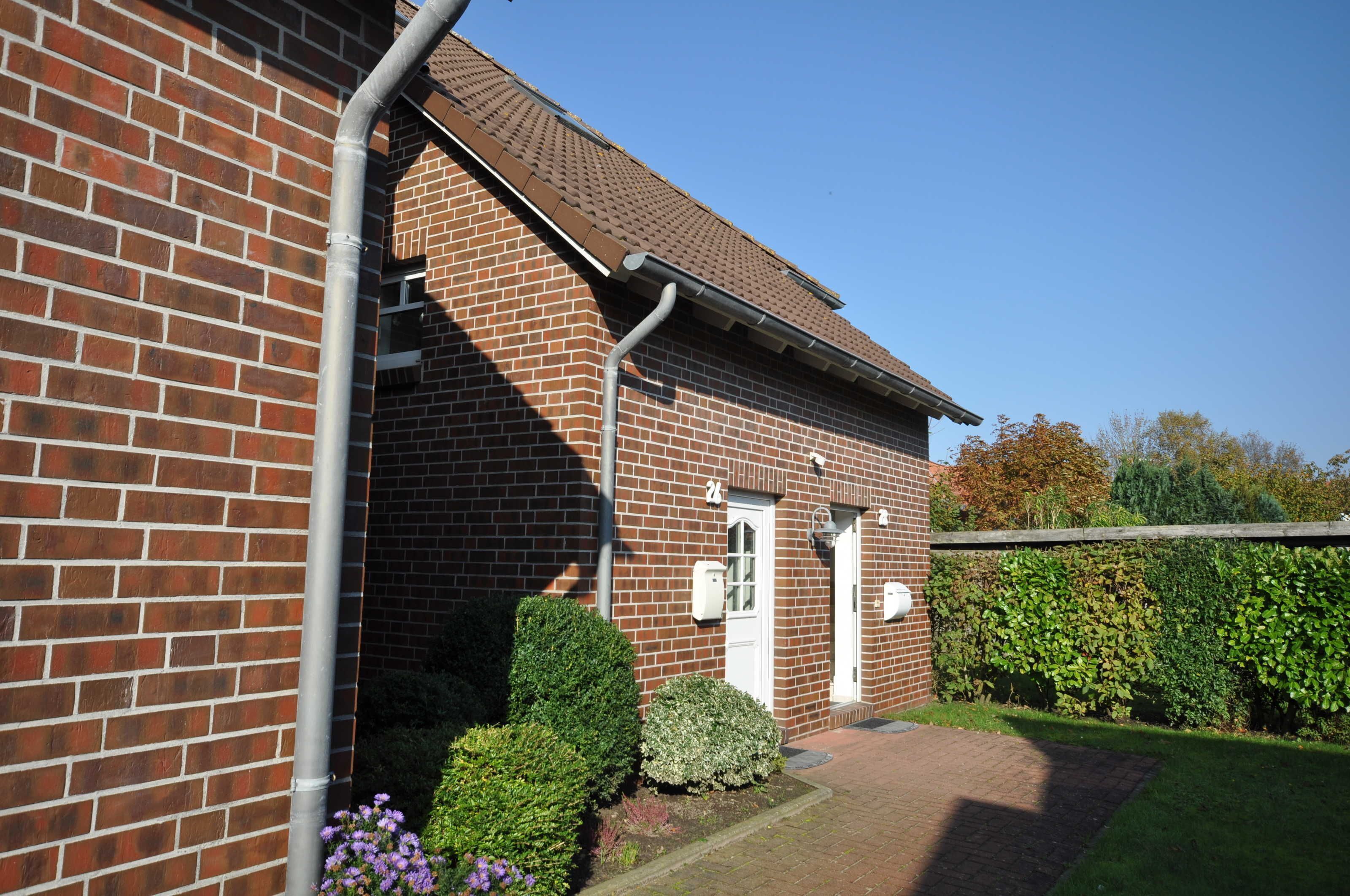 A brick building with a tiled roof, a white door, and a small garden area with hedges and flowers.