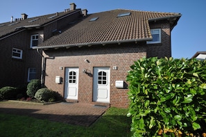 Une maison à deux étages, construite en briques, avec un toit en tuiles, des portes numérotées et un petit jardin.