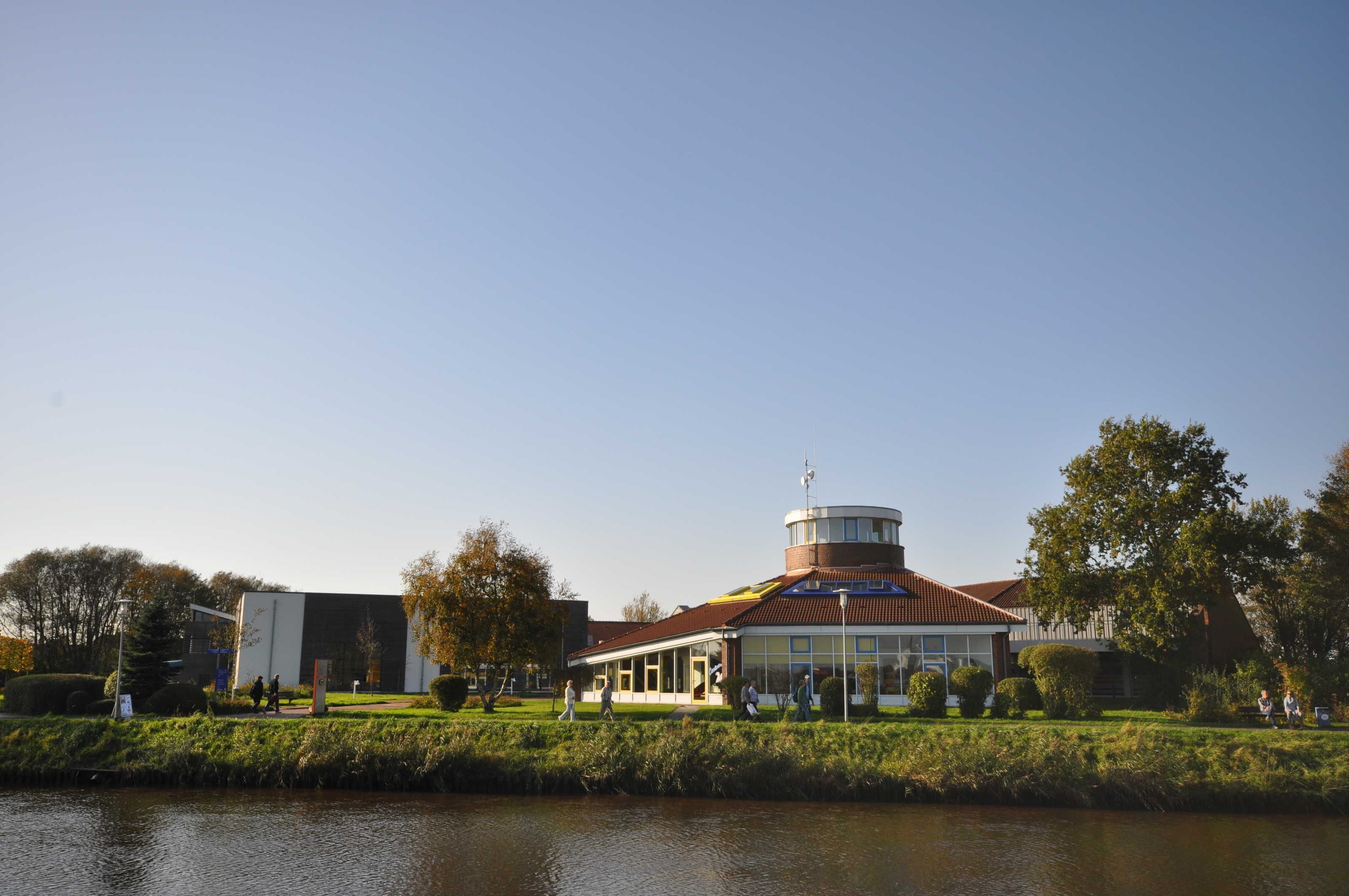 A building with a circular tower, a flat-roofed structure, and a water body in the foreground.