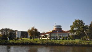 A building with a circular tower, a flat-roofed structure, and a water body in the foreground.